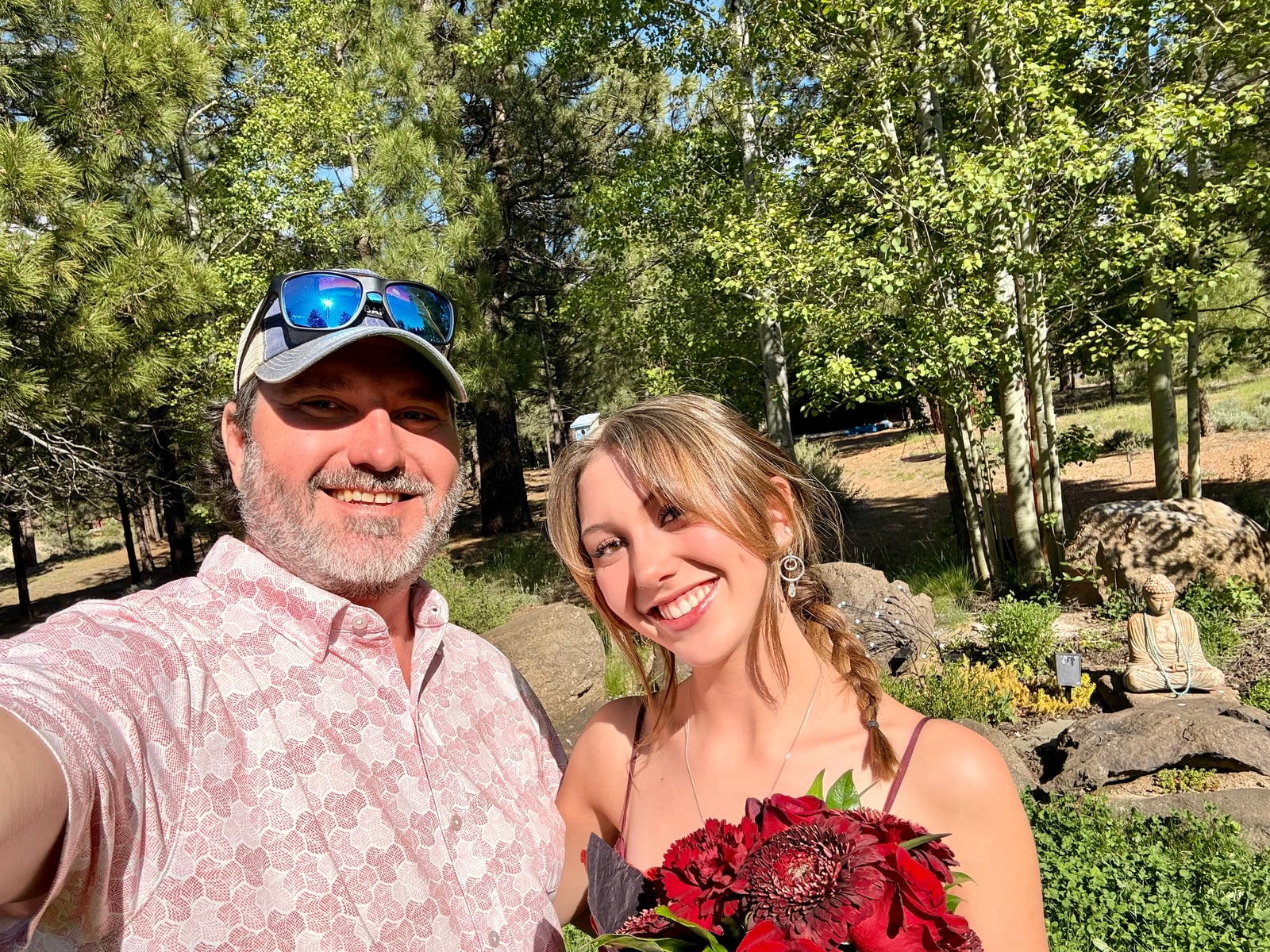Two smiling people take a selfie outdoors, one holding a red flower bouquet, with trees and rocks in the sunny background.