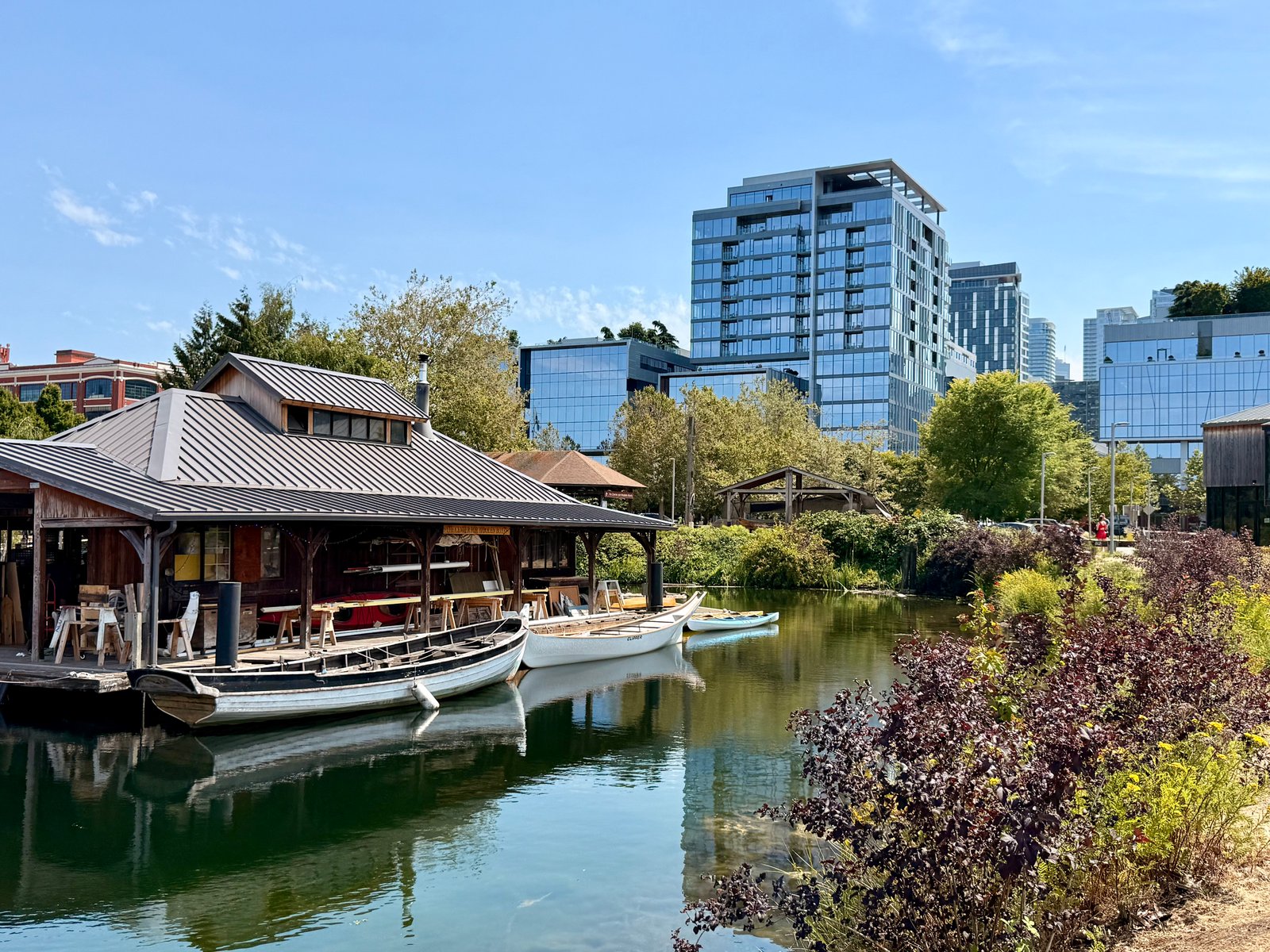 A wooden boathouse with boats on calm water, surrounded by foliage, with modern glass buildings in the background.