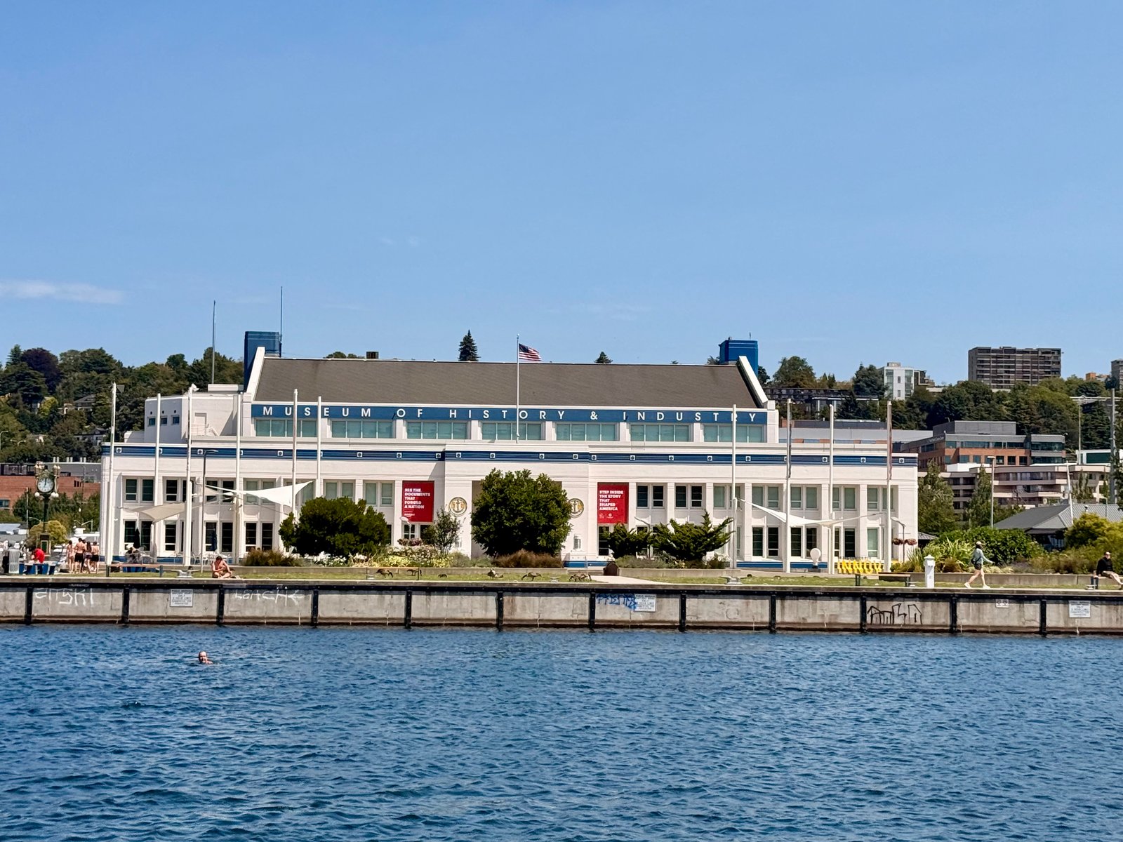 The Museum of History & Industry building stands beside blue water with people on its shore.