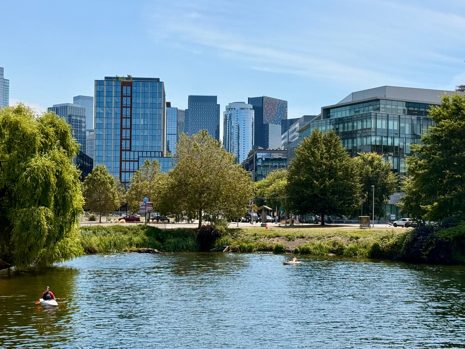 City skyline visible behind a tree-lined bank and a body of water with two people on small boats.