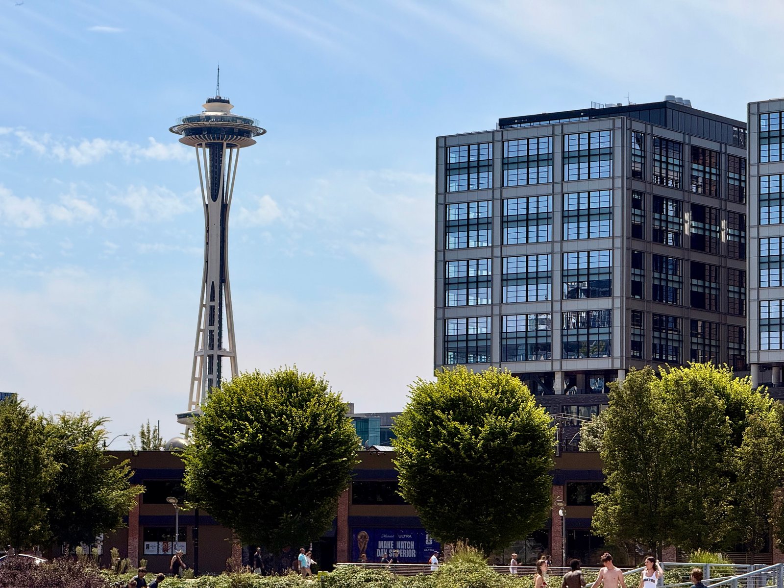The Space Needle and a modern building stand against a blue sky, with green trees and people in the foreground.