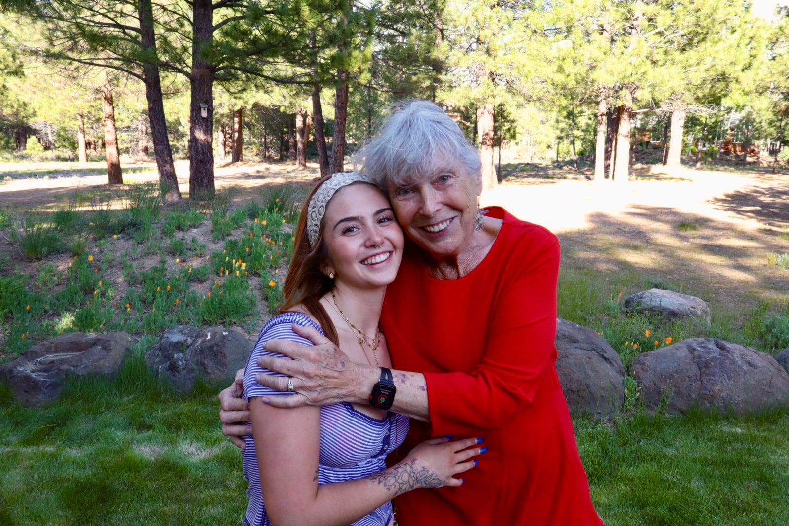 Two people hug and smile outdoors in a grassy area with trees, rocks, and orange flowers.