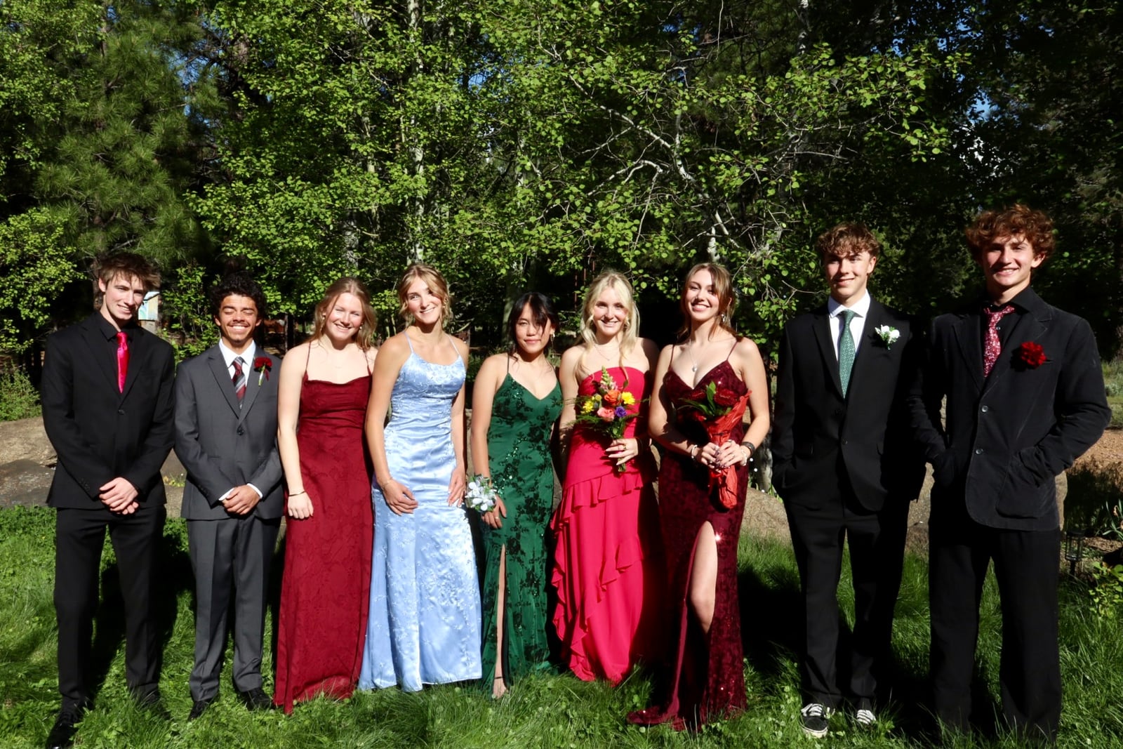 Nine young people dressed in formal attire stand outdoors on grass with green trees in the background.