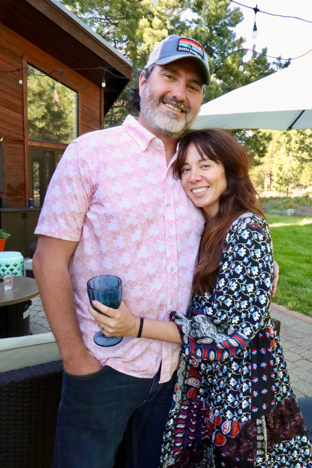 Two smiling people, a man with a beard and a woman holding a blue glass, embracing outdoors with a house and trees.