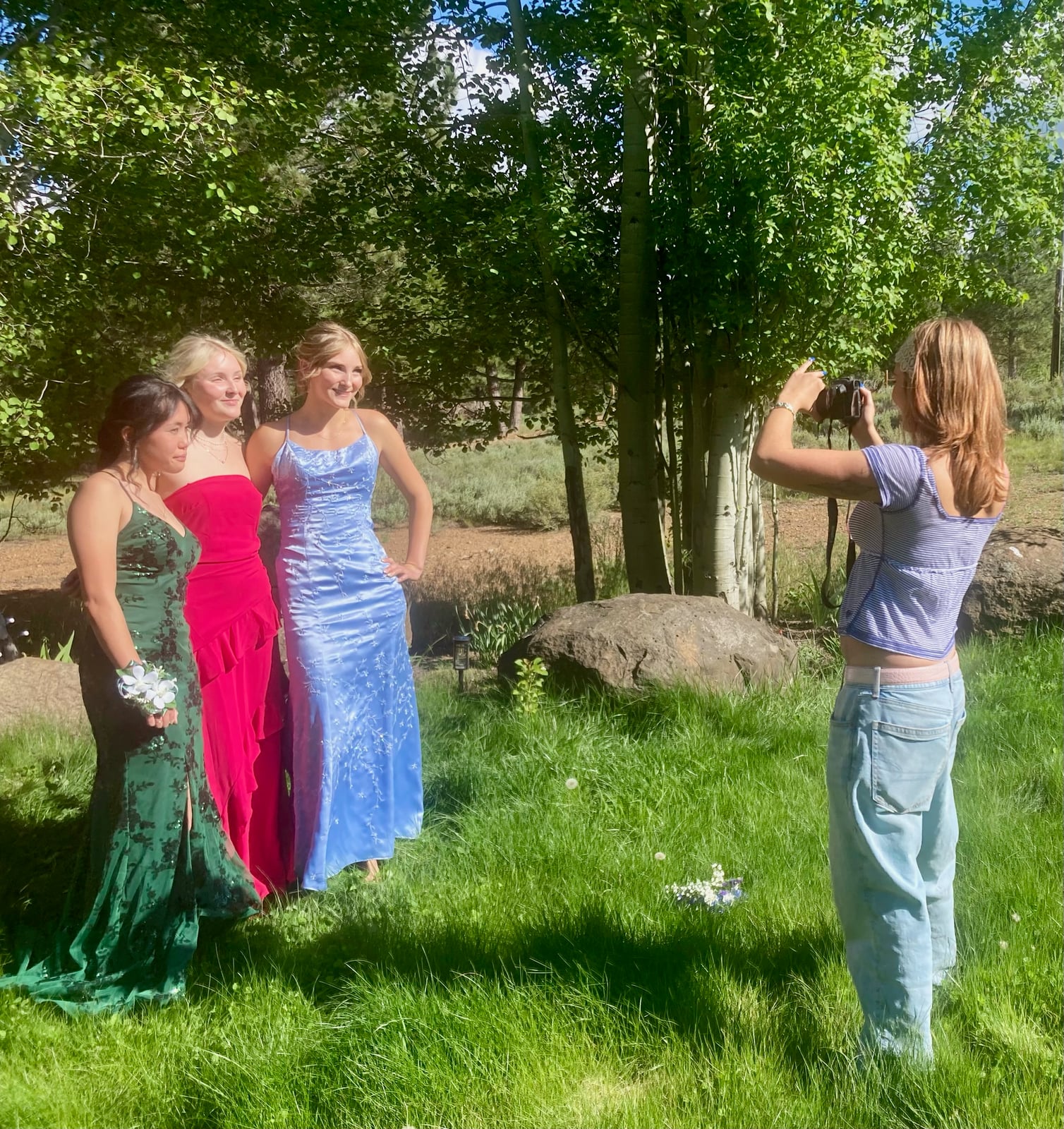 Three young women in formal dresses pose while another person photographs them in a bright, grassy, wooded area.
