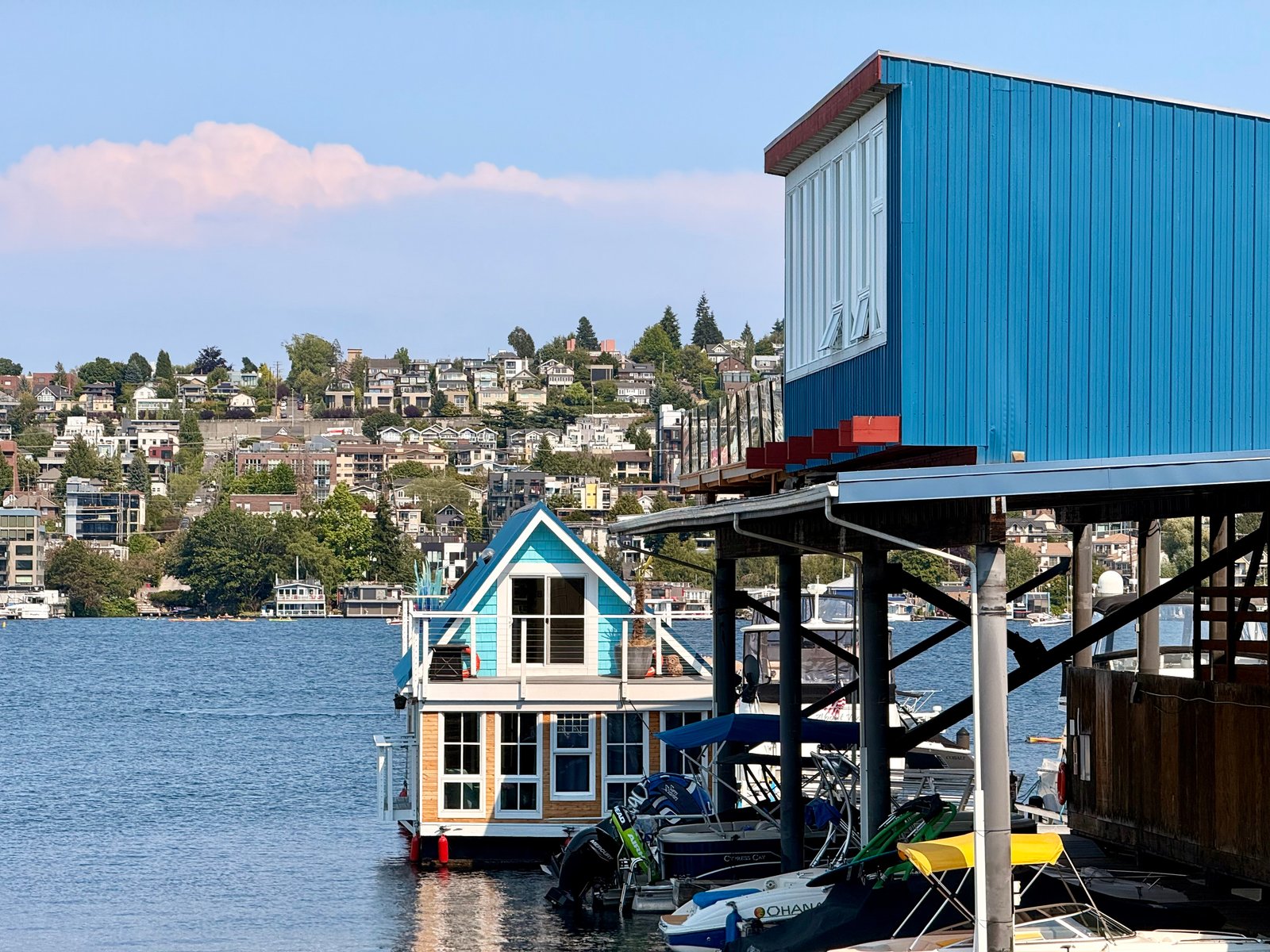 Boathouses and docked boats on a lake, with a hillside town of houses and trees in the background.